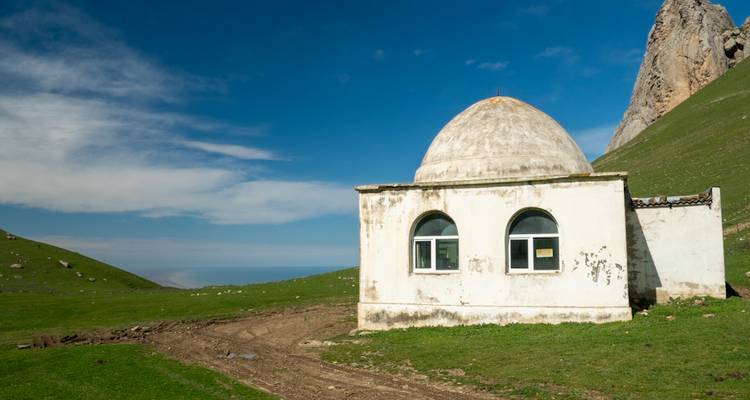 Un petit sanctuaire blanc aux dômes arrondis se dresse seul sur une prairie alpine verdoyante en contrebas des pics rocheux.