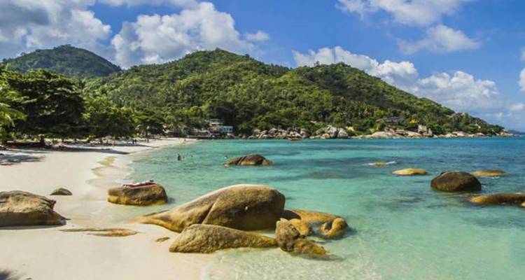 Playa tropical idílica con arena blanca, agua turquesa, rocas de granito y colinas boscosas.