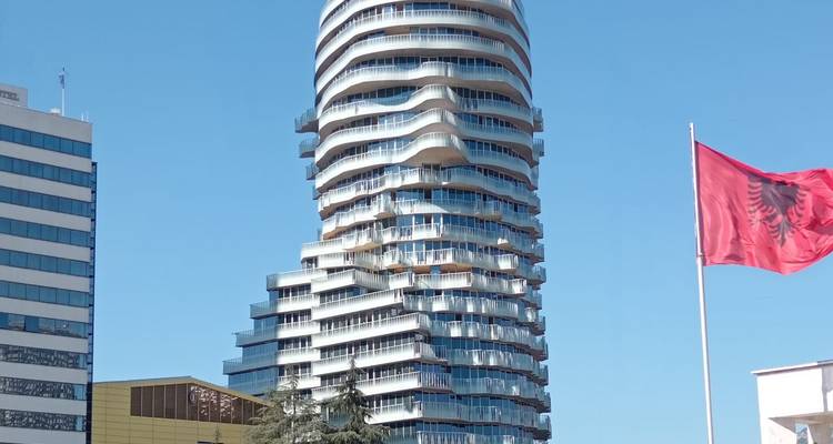 Modern twisting high-rise tower with balconies beside an Albanian flag against a clear sky