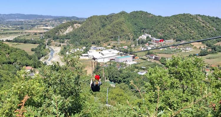 Zipliner wearing red helmet gliding above a green valley dotted with buildings