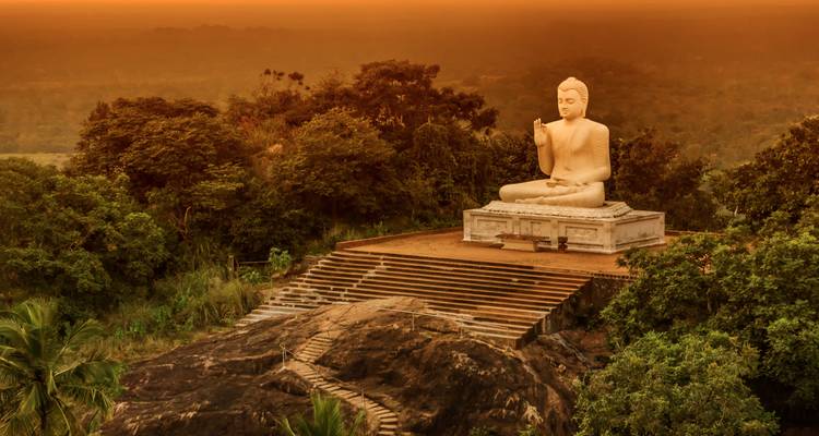 Grande statue de Bouddha en position assise sur une plateforme au sommet d'une colline, au-dessus de la jungle, lors d'un coucher de soleil dramatique aux teintes orangées.