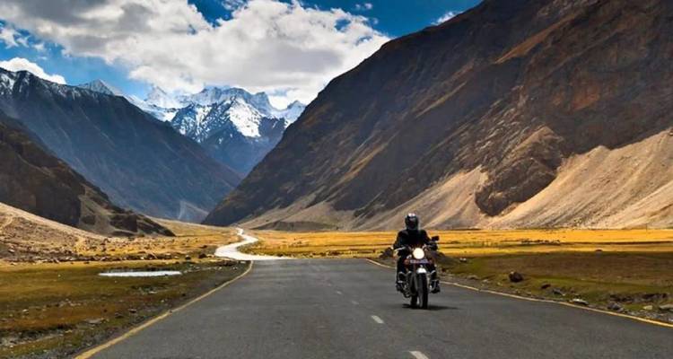 Einsamer Motorradfahrer fährt auf einer leeren Asphaltstraße durch ein dramatisches Himalaya-Tal, das von zerklüfteten Gipfeln umrahmt wird.