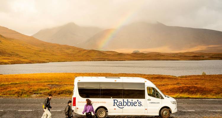 Rondritminibus met bedrijfslogo geparkeerd aan een loch met een regenboog die boven mistige Highlands-heuvels boven uit steekt