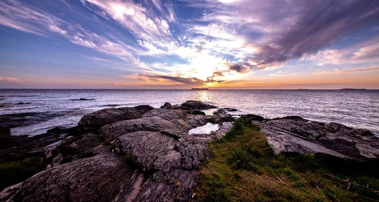 Dramatischer Sonnenuntergang über dem Ozean malt den Himmel, während Wellen gegen zerklüftete Küstenfelsen krachen.