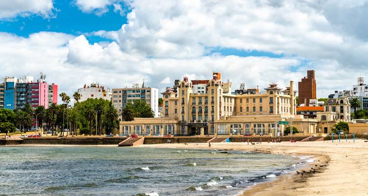 Historisches strandnahes casinoähnliches Gebäude mit Blick auf sandigen Strand und sanfte Wellen mit Stadtskyline im Hintergrund.