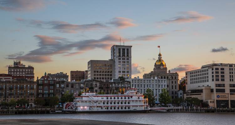 Savannah riverfront skyline at sunset with paddlewheel riverboat docked in foreground.