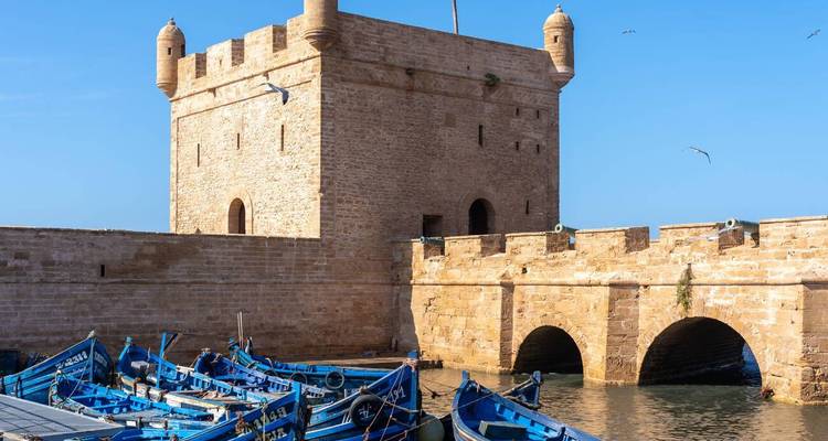 Fort et pont en pierre historiques surplombant des bateaux de pêche bleus dans le port d'Essaouira.