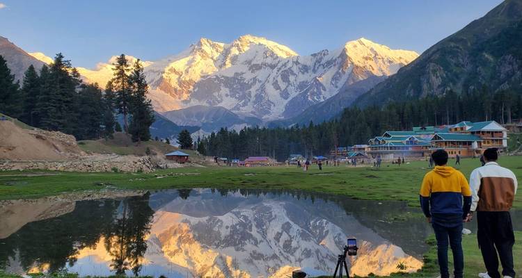 Deux jeunes voyageurs admirent un panorama montagneux lumineux reflété dans un étang calme au crépuscule.
