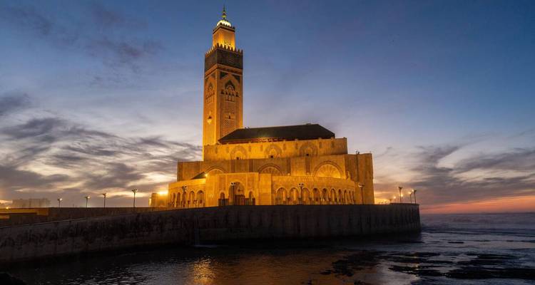 Mosquée Hassan II illuminée brillant d'or au coucher du soleil au bord de l'océan Atlantique sous un ciel dramatique de crépuscule