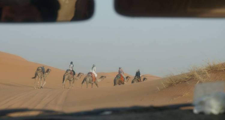 Vue à travers un pare-brise de véhicule d'une petite caravane de chameaux traversant un terrain désertique sablonneux au loin