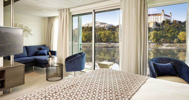 Geräumige Kreuzfahrtkabine mit Balkonblick auf eine Burganlage auf dem Hügel gegenüber am Fluss