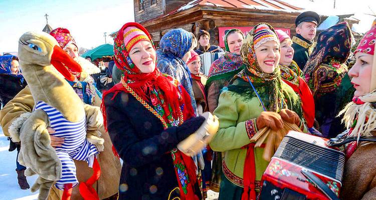 Mujeres vestidas festivamente con bufandas coloridas celebran al aire libre en la nieve invernal, sosteniendo instrumentos y una cigüeña de peluche.