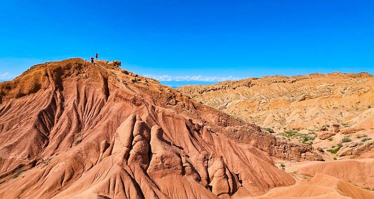 Crête de canyon sculptée rouge et orange avec deux randonneurs debout au sommet sous un ciel bleu éclatant.