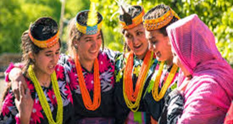Smiling Kalash women in vibrant traditional dresses and beaded headdresses huddle together outdoors.