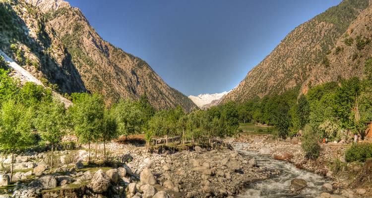 Panoramic alpine valley with rushing river, stone slopes and snow-capped peaks in the distance under clear sky.