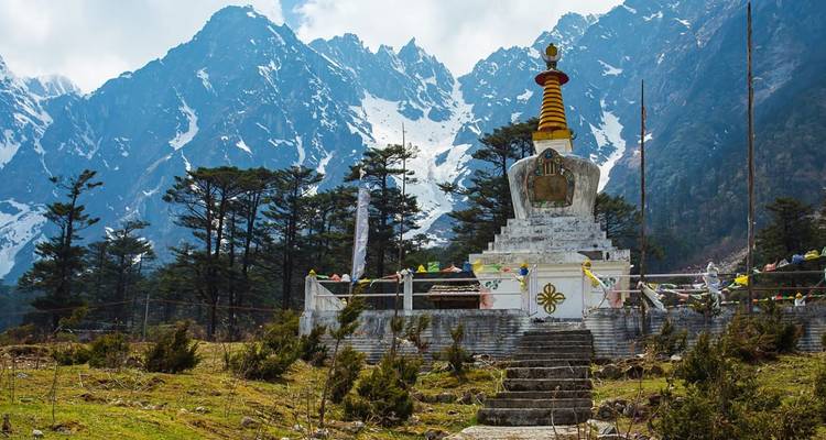 Weiße buddhistische Stupa mit Gebetsfahnen behängt vor hohen Kiefern und dramatischen schneebedeckten Himalaya-Gipfeln.