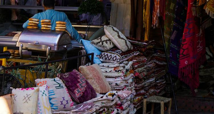 Stapel von gewebten Teppichen und Kissen in einem lebendigen Souk-Stand mit einem Verkäufer, der in der Nähe Sandwiches grillt