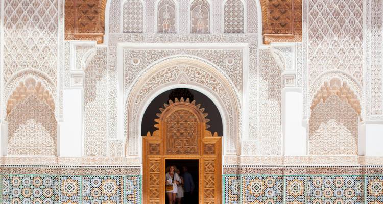 Aufwendig geschnitzter und gefliester Eingang einer marokkanischen Madrasa mit kunstvollen Stuckarbeiten und Holztür, zwei Besucher stehen im Inneren