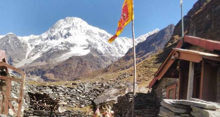 Snow-capped Himalayan peak rises behind rocky stone huts and a fluttering prayer flag on a sunny day