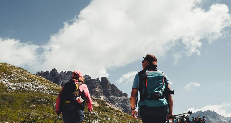 Two hikers with backpacks and trekking poles walking toward jagged mountain peaks under large cloud