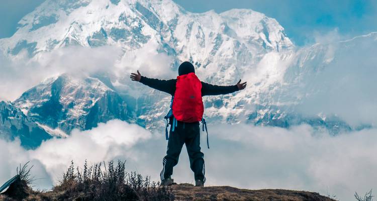 Solo trekker with red backpack raises arms facing towering snow-covered Himalayan massif amid clouds