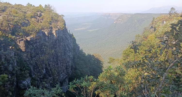 El borde rocoso del acantilado desciende hacia un vasto valle verde con cordilleras distantes y brumosas.
