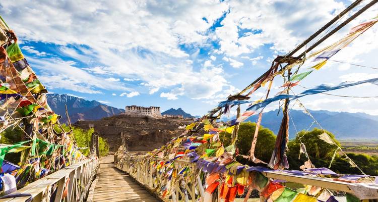 Mit Gebetsfahnen geschmückte Holzbrücke, die zu einem entfernten Kloster unter hellem Himmel führt.