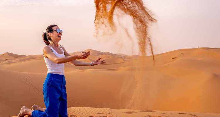 Femme souriante agenouillée dans les dunes lançant un panache de sable haut dans l'air du désert