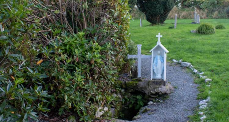 Pozo santo rural y tranquilo con un pequeño santuario de la Virgen María y una cruz de piedra junto a un camino de grava en un cementerio cubierto de hierba.