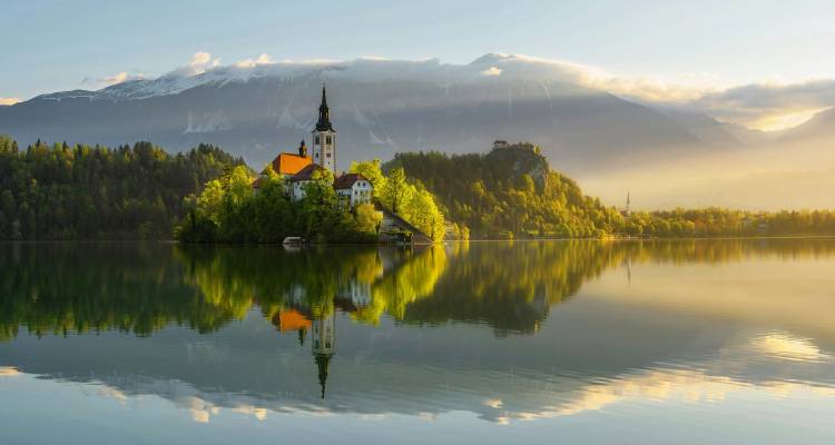 Fairytale Church of the Assumption on Lake Bled isle mirrored perfectly in still water with snowcapped Alps.