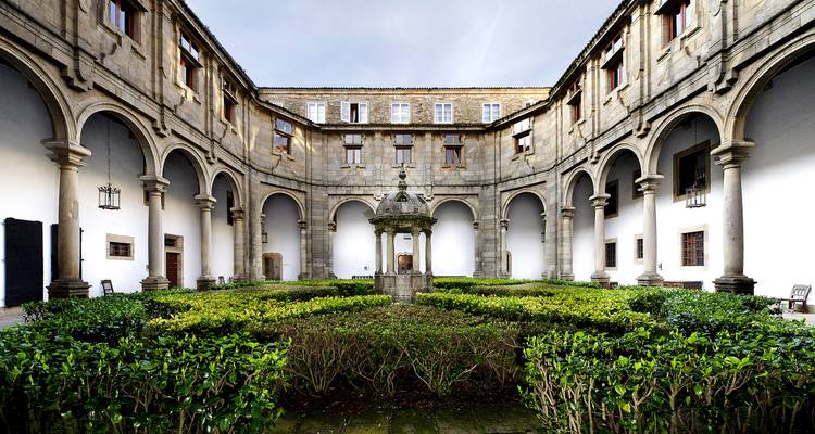 Claustro histórico de piedra con columnatas arqueadas, setos recortados y una fuente decorativa bajo el cielo abierto.