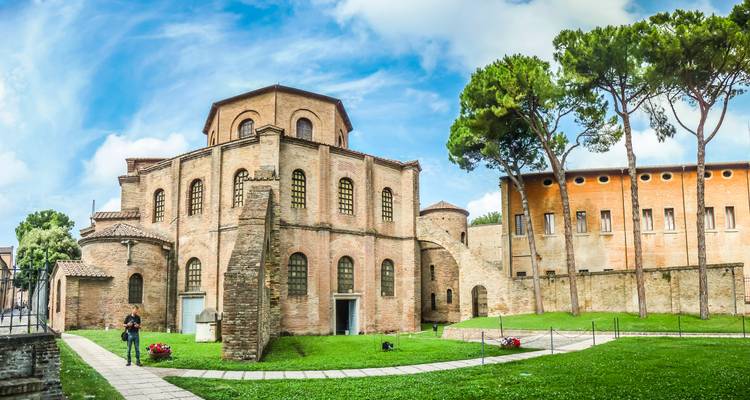 Panoramablick auf die achteckige Basilika San Vitale, umgeben von grünen Rasenflächen und hohen Kiefern in Ravenna.