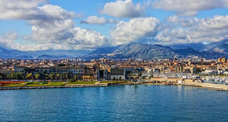 Panoramisch uitzicht op Palermo's waterkant met een jachthaven, historische gebouwen en bergen onder gedeeltelijk bewolkte blauwe luchten