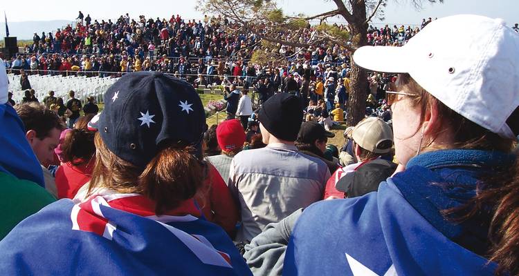 Gran multitud envuelta en banderas australianas asistiendo a una ceremonia Anzac al aire libre con vista al mar.