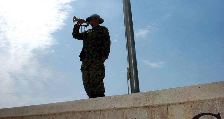 Silhouetted bugler in military uniform sounding a call against an open blue sky.