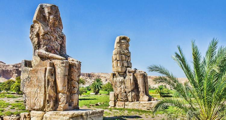 The twin Colossi of Memnon statues rise above green fields under a clear desert sky.