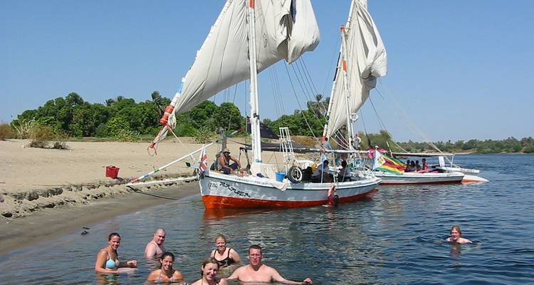 Felucca sailboat anchored by a sandy Nile bank while swimmers enjoy the refreshing water.