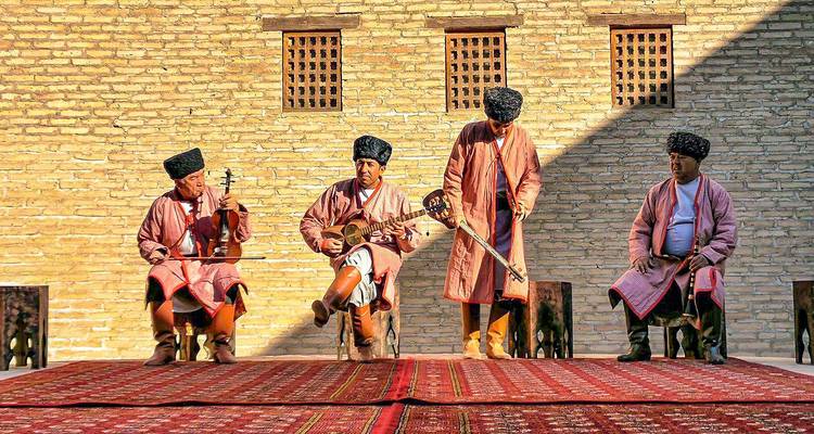 Musiciens traditionnels en tenue régionale jouant des instruments à cordes sur une scène recouverte de tapis avec un fond de briques.