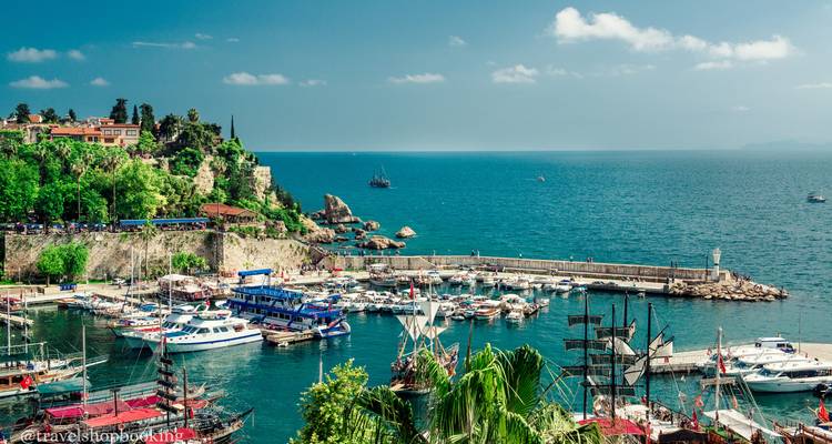 Un puerto deportivo vibrante bordeado de embarcaciones y muros históricos se abre al mar turquesa de Antalya en un día soleado.
