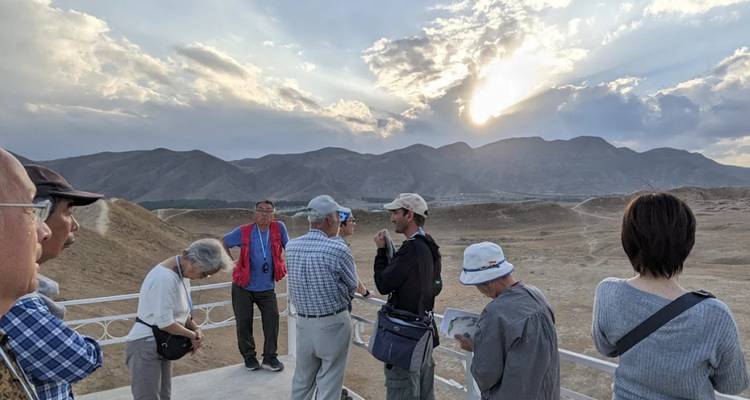 Los viajeros se encuentran en una plataforma de observación con vista a colinas áridas mientras el sol se filtra a través de las nubes.