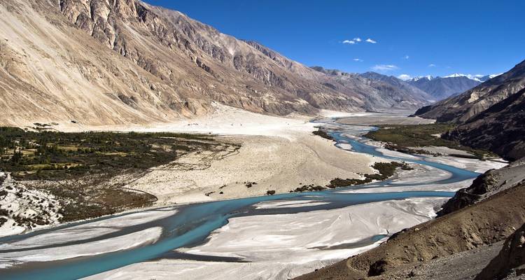 Weite Luftaufnahme eines geflochtenen türkisfarbenen Flusses, der sich durch ein weites trockenes Tal schlängelt, umgeben von kahlen Gipfeln unter einem tiefblauen Himmel.