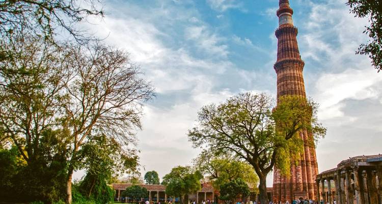 Der hohe rote Sandstein-Qutub-Minar-Turm erhebt sich über belaubte Bäume mit Besuchern, die sich am Fuß unter einem blauen, teilweise bewölkten Himmel tummeln.