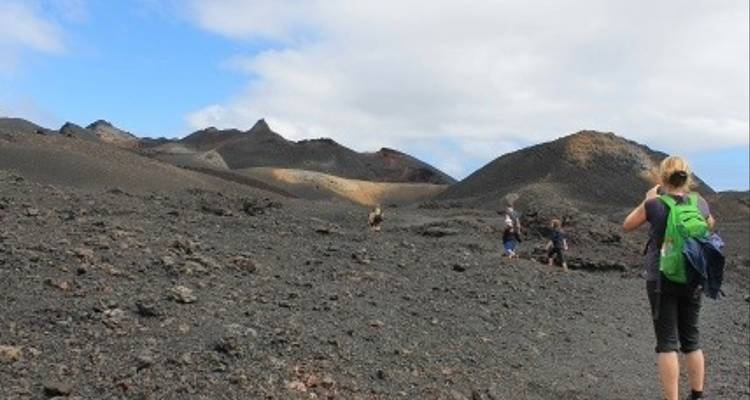 Wandelaars doorkruisen kaal zwart vulkanisch landschap onder gedeeltelijk bewolkte hemel.