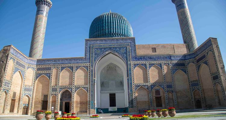 Côté cour du complexe de Gur-e Amir avec des parterres de fleurs colorés sous un ciel bleu dégagé.