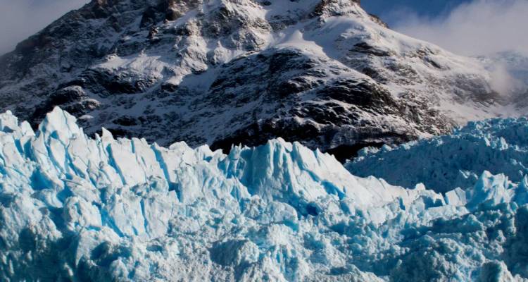 Pared de glaciar azul dentado que se eleva bajo montañas cubiertas de nieve bajo cielos melancólicos.