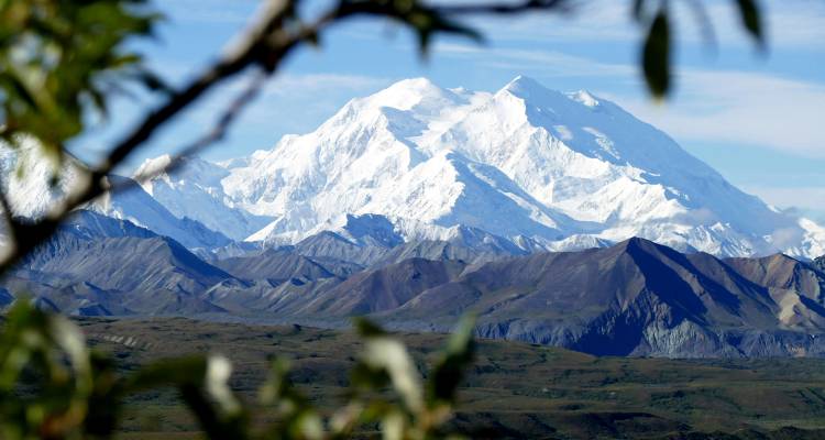 Snow-capped Mount Denali dominates the horizon above rugged ridges and a green Alaskan valley.