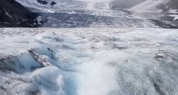 Superficie azul hielo de un glaciar gigantesco con grietas profundas que se extienden hacia picos lejanos.