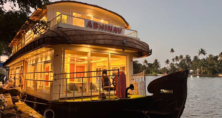 A warmly lit luxury houseboat named "ABHINAV" is moored on tranquil backwaters at dusk.