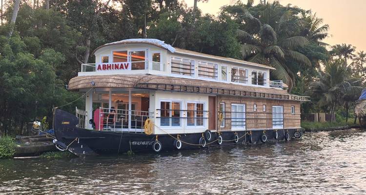 A large wooden houseboat with upper deck windows floats beside lush palms on calm waters.