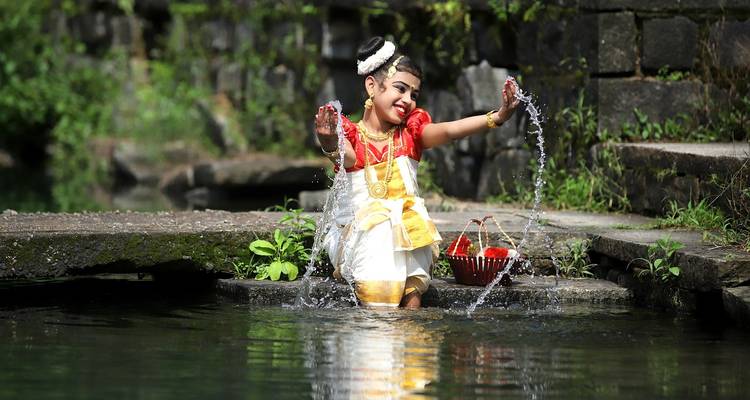 A young girl in traditional Kerala costume splashes water joyfully in a forest pool.